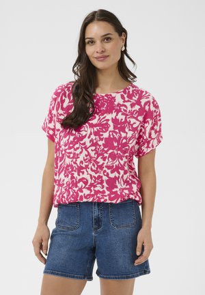 Žena s dlhými tmavými vlasmi, oblečená v ružovo-bielom kvetovanom tričku s krátkym rukávom a modrých džínsových šortkách, stojaca pred jednoduchým pozadím.