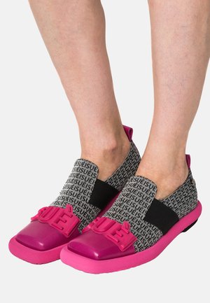 Piedi che indossano scarpe slip-on nere e grigie con suole e punte rosa, con la parola "USUE" in lettere rosa in rilievo sulla parte superiore.