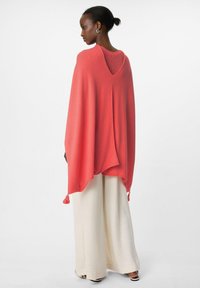 Koronkowy poncho w kolorze koralowym z głębokim cięciem na plecach, z frędzlami na dolnym brzegu. Połączony z luźnymi, kremowymi spodniami typu wide-leg.