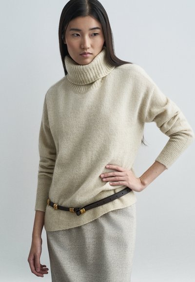 Pull à col roulé beige en tissu tricoté doux, avec un col et des poignets côtelés. Associé à une jupe gris clair et une ceinture marron avec des accents dorés.