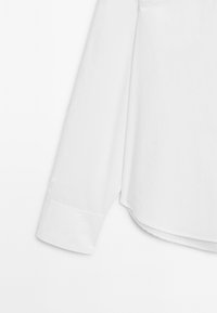 Polsino e parte inferiore della manica della camicia bianca a maniche lunghe con orlo abbottonato, su sfondo bianco uniforme.