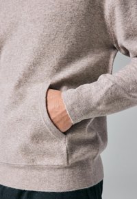 Hellbraun Sweatshirt mit einer weichen Textur, ausgestattet mit einer Kängurutasche und gerippten Bündchen an den Handgelenken und am Saum. Nahaufnahme.