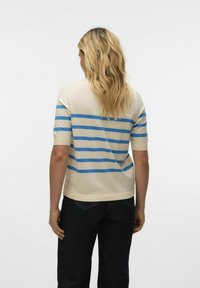 Gebreide trui in crèmekleur met blauwe horizontale strepen, korte mouwen en een ronde halslijn. Gedragen met donkere spijkerbroek, gezien vanaf de achterkant.