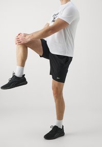 Černé sportovní šortky v kombinaci s bílým tričkem s krátkým rukávem. Boty jsou černé s bílými akcenty a model je v dynamické póze.