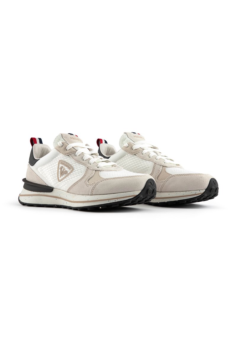 Beige and white athletic shoes with mesh and suede textures, black soles, and a logo on the side; features red and blue accents at the heel.