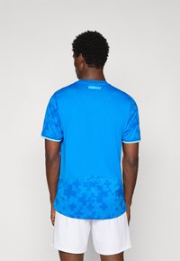 Man wearing a bright blue sports shirt with geometric patterns on lower back and sleeves, paired with plain white shorts, facing away.