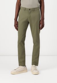 Pantalones de algodón verde oliva con un corte ajustado, que presentan un cierre de botón frontal y bolsillos, combinados con zapatillas de color claro.
