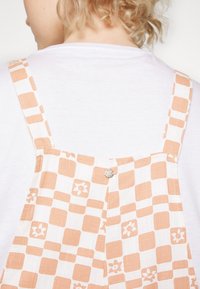 Bretelles de salopette en coton blanc avec un motif géométrique pêche, dotées d'un bouton argenté et d'une finition douce et texturée.