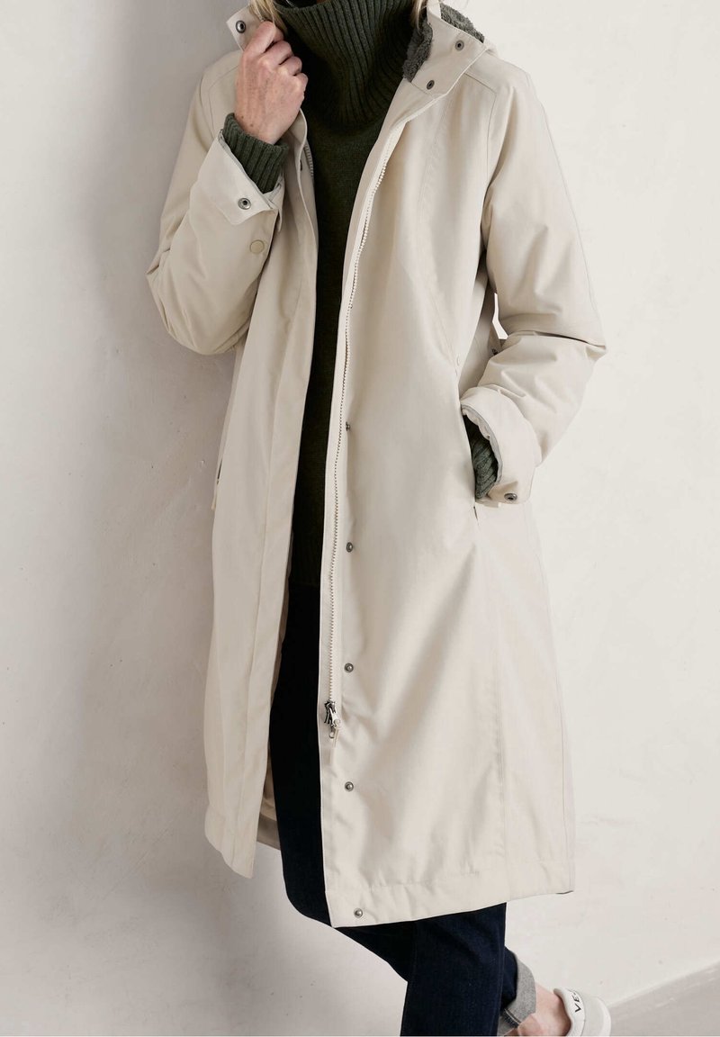 Cappotto impermeabile beige con chiusura a zip e bottoni automatici, dotato di cappuccio e tasche laterali, indossato sopra un maglione verde scuro e jeans scuri.
