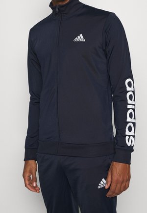 Man draagt een zwart Adidas trainingspak met wit logo op de borst, linkermouw en linkerbeen.