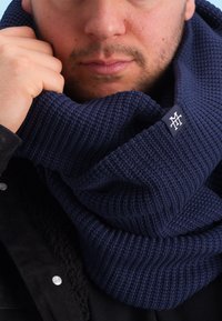 Navy knitted scarf with a ribbed texture and folded design, featuring a small white logo tag. Worn snugly around the neck.