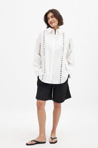 Camicia bianca con dettagli ricamati lungo i bordi, indossata con pantaloni corti neri e infradito. La camicia ha una vestibilità rilassata e chiusura con bottoni.