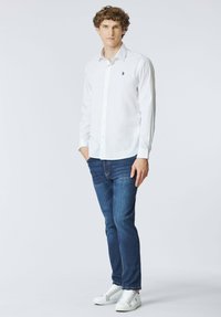 Giovane uomo con capelli ricci che indossa una camicia bianca con bottoni, jeans blu e sneakers bianche, in piedi contro uno sfondo chiaro e semplice.