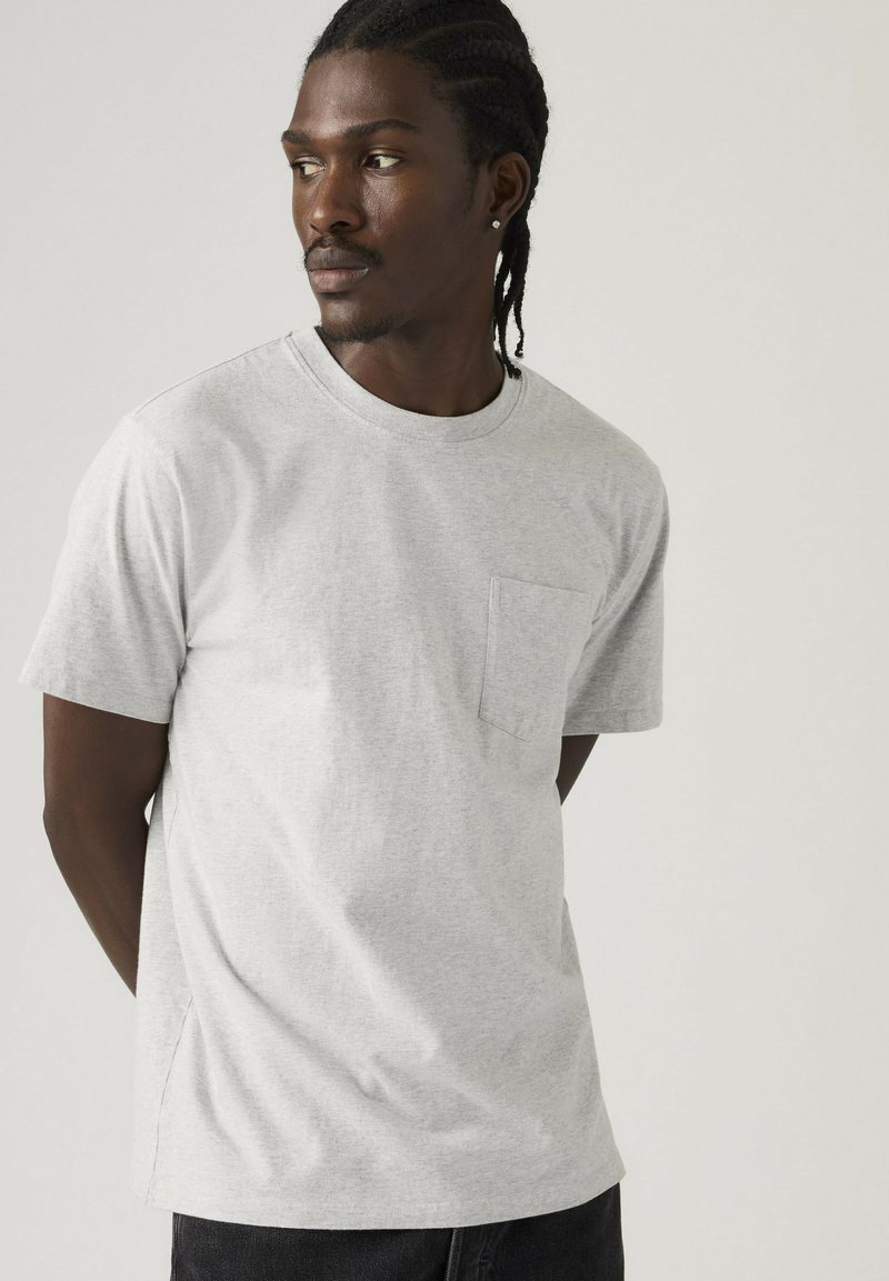 Mann mit geflochtenem Haar trägt ein hellgraues kurzärmeliges T-Shirt mit Brusttasche, schaut nach links vor einem schlichten Hintergrund.