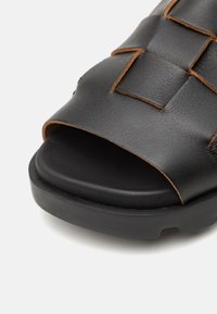 Infradito in pelle nera con design superiore intrecciato, punta aperta e cuciture marroni a contrasto su una robusta suola nera.