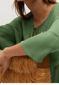 Blusa verde a trama con maniche ampie, caratterizzata da un dettaglio a fiocco sul collo. Una borsa intrecciata è tenuta in una mano accanto alla blusa.