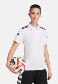Witte polo met zwarte accenten, korte mouwen en een drie-strepen ontwerp. Een voetbal wordt in één hand vastgehouden, met een zwarte short eronder.
