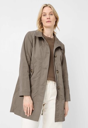 Frau trägt einen hellbraunen, geknöpften Trenchcoat über einem braunen Strickpullover und weißer Hose, steht vor einem weißen Hintergrund.