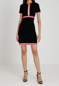 Femme portant une robe noire à manches courtes avec des rayures rouges et blanches sur les manches, à la taille, à l'ourlet et une fermeture éclair verticale à l'avant, associée à des talons noirs.