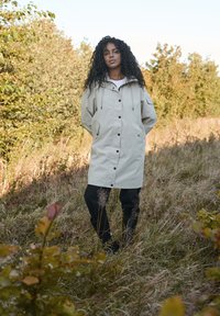 Beige wasserfester Mantel mit Kapuze, Knopfverschluss vorne und seitlichen Taschen. Darunter werden schwarze Hosen getragen. Natürliche Umgebung im Freien mit Gras.