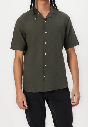 Homme portant une chemise boutonnée à manches courtes vert foncé avec un pantalon noir, debout devant un fond blanc uni.