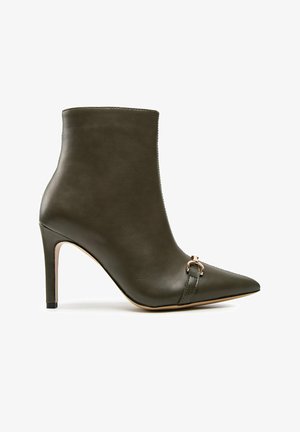 Tmavě zelená kožená kotníková bota na vysokém podpatku s ostrou špičkou a dekorativní kovovou přezkou na přední straně.