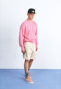 Rosa Sweatshirt, beige Shorts, rosa Sneakers mit weißen Akzenten, schwarze Cap mit Logo. Stehend auf blauem Boden vor schlichtem Hintergrund.
