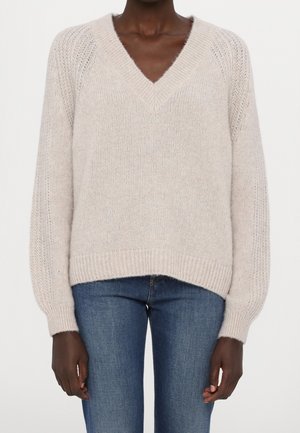 Personne à la peau foncée portant un pull en maille beige clair à col en V et un jean en denim bleu, debout devant un fond uni.