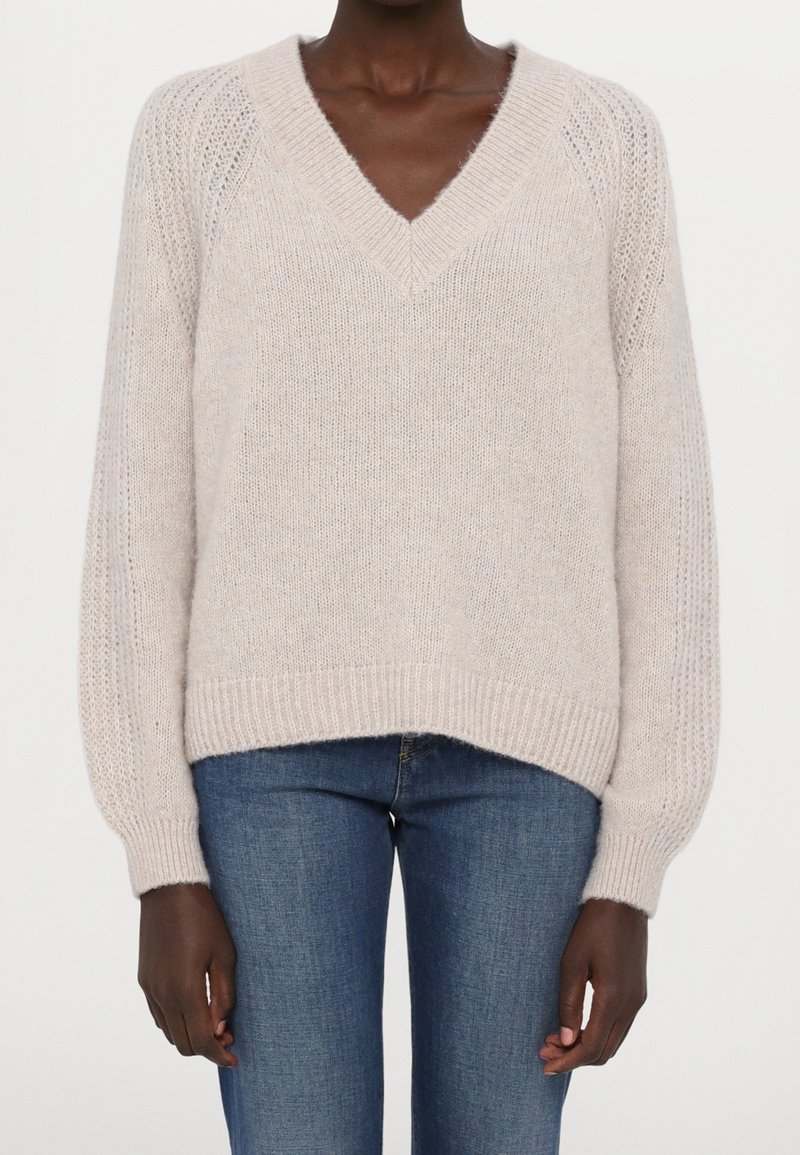 Personne à la peau foncée portant un pull en maille beige clair à col en V et un jean en denim bleu, debout devant un fond uni.