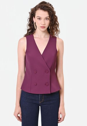 Donna con capelli ricci che indossa orecchini a cerchio dorati, un top viola senza maniche doppiopetto e jeans blu scuro su sfondo bianco.