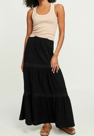 Femme portant un haut beige sans manches côtelé et une longue jupe noire à volants avec des sandales marron, debout devant un fond clair uni.