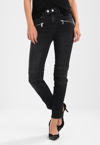 Jeans skinny negros de cintura alta, con bolsillos frontales con cremallera y detalles en los paneles de las rodillas. La tela tiene un acabado suave y ligeramente desgastado.