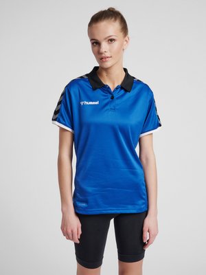 Ung kvinde iført en blå Hummel sportspolo med sort krave og sorte cykelshorts står mod en ensfarvet baggrund.