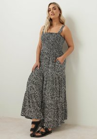 Svart och vit mönstrad maxiklänning med smockad byst, fyrkantig halsringning, justerbara axelremmar och fickor, i kombination med svarta sandaler.