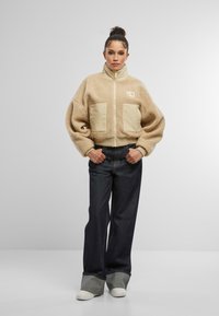 Giacca in pile beige con colletto alto, chiusura con zip e due tasche frontali. Abbinata a pantaloni in denim scuri a gamba larga e sneaker bianche.