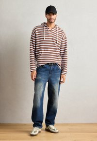 Stripet brun og hvit hettegenser med snorer, parret med falmede blå jeans og grå og grønne sneakers, mot en nøytral bakgrunn.