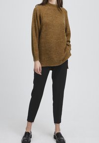Mujer vestida con un suéter de punto marrón, combinado con pantalones negros entallados y mocasines negros, de pie frente a un fondo claro y liso.