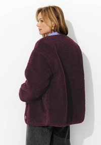 Veste en fausse fourrure bordeaux à coupe décontractée, avec un détail de bouton, un col rond et une surface texturée. Portée par-dessus un pantalon sombre.