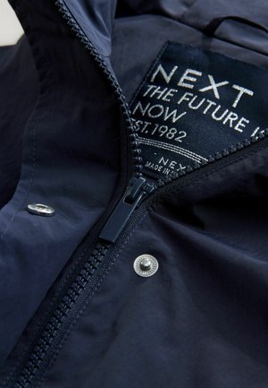Mörkblå jacka med delvis öppen dragkedja, som visar en etikett med vit text "NEXT THE FUTURE NOW EST. 1982", metallknappar synliga.