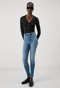 Black long-sleeve bodysuit with buttons, paired with light blue high-waisted skinny jeans featuring distressed details. Black loafers complete the look.