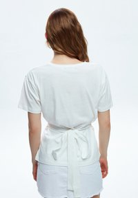 White cotton t-shirt with short sleeves, featuring a tie-back detail at the waist and a relaxed fit. Smooth texture, simple design.