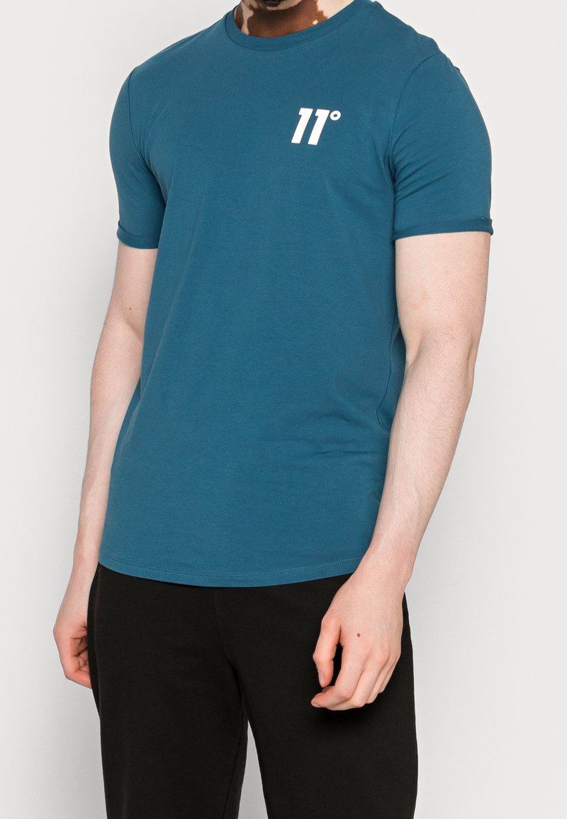 Man som bär en åtsittande kortärmad t-shirt i teal med vitt "11°"-logotyp på bröstet, i kombination med svarta byxor mot en neutral bakgrund.