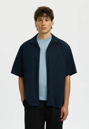 Jauns vīrietis ar cirtainiem matiem, valkājot gaiši zilu apaļkakla kreklu zem īsrokainas melnas pogām aizpogātas krekla, stāv ar rokām kabatās.