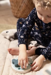 Houten puzzel met kleurrijke vormen, waaronder een blauwgroen hondenbot en een witte cilinder, op een lichte quilt. Hand van een kind die een stuk vasthoudt.