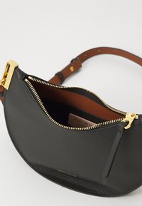 Sac en cuir noir avec un design incurvé, des fermetures éclair dorées et un intérieur marron. Il possède une bandoulière amovible et une petite étiquette de logo à l'intérieur.