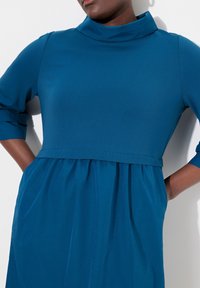 Sininen, pitkähihainen mekko korkealla kaula-aukolla, muotoonommeltu vartalohame ja levenevä helma. Pehmeä tekstuuri, sivutaskut ja rento istuvuus.