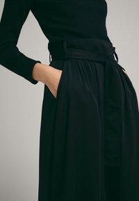 Femme portant un haut noir à manches longues et une jupe noire taille haute avec une large ceinture, les mains posées négligemment dans les poches avant.