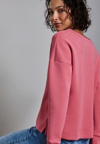 Roze sweatshirt met lange mouwen, een ronde hals, ontspannen pasvorm, zijsplitten aan de zoom en een gladde textuur. G gedragen met denim jeans.