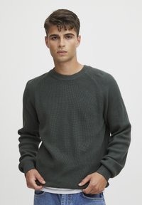 Pulover tricotat de culoare verde închis, cu textură ribaltă, guler rotund și mâneci raglan. Purtați peste o cămașă albă, asortat cu blugi de denim ușor albastru.