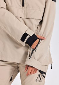 Beige waterdichte jas met verstelbare manchetten, zwarte binnenvoering en logo-accent. Inclusief ritszakken en versterkte naden.